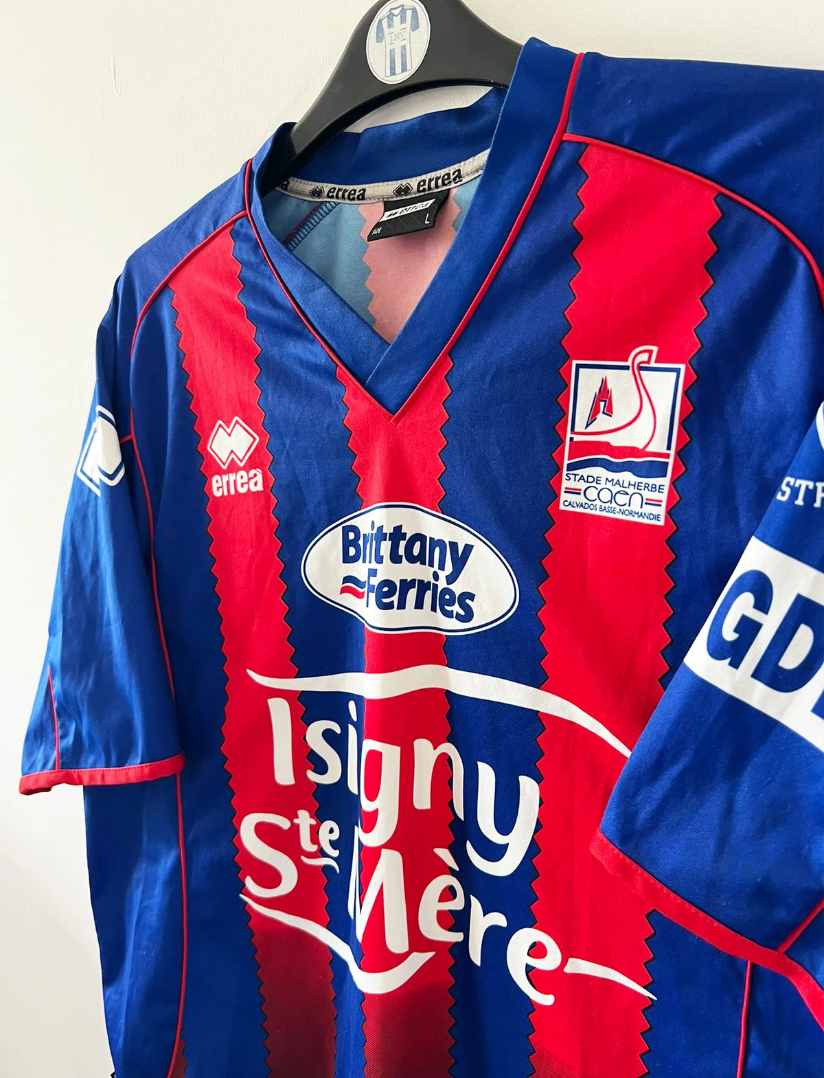Maillot de foot vintage rouge et bleu du SM Caen de la saison 2004/2005. Il s'agit du maillot édité pour la finale de la Coupe de la Ligue. On peut retrouver les sponsors Isigny Ste Mère et Brittany Ferries. Il s'agit d'un maillot authentique d'époque.