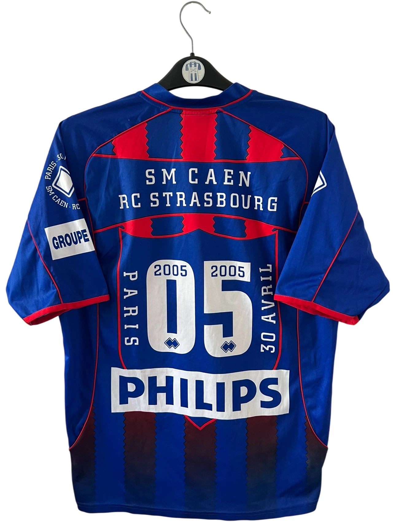 Maillot de foot vintage rouge et bleu du SM Caen de la saison 2004/2005. Il s'agit du maillot édité pour la finale de la Coupe de la Ligue. On peut retrouver les sponsors Isigny Ste Mère et Brittany Ferries. Il s'agit d'un maillot authentique d'époque.
