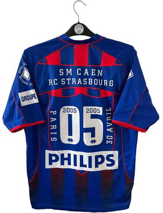 Maillot de foot vintage rouge et bleu du SM Caen de la saison 2004/2005. Il s'agit du maillot édité pour la finale de la Coupe de la Ligue. On peut retrouver les sponsors Isigny Ste Mère et Brittany Ferries. Il s'agit d'un maillot authentique d'époque.