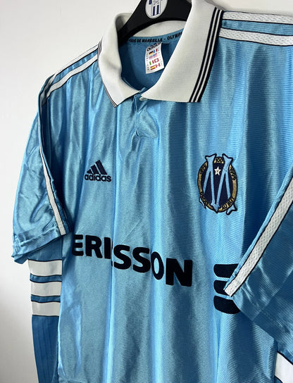 Maillot de foot vintage extérieur bleu de l'Olympique de Marseille de la saison 1998/1999. On peut retrouver l'équipementier Adidas et le sponsor Ericsson. Il s'agit d'un maillot authentique d'époque.
