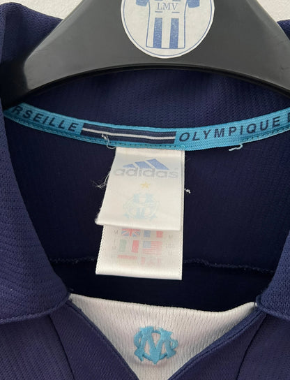 Maillot de foot vintage OM 1999/2000 extérieur bleu. On peut retrouver l'équipementier Adidas et le sponsor Ericsson. Il s'agit d'un maillot authentique d'époque.