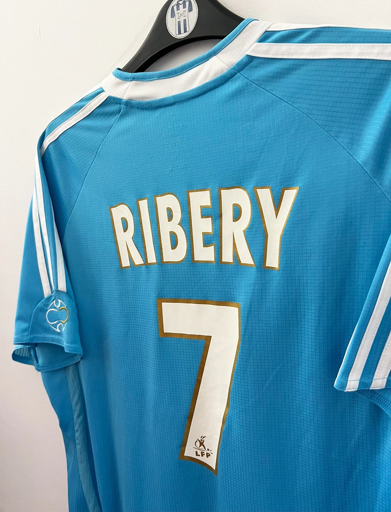 Maillot de foot vintage extérieur bleu et blanc de l'OM 2006/2007. On peut retrouver l'équipementier Adidas et le sponsor Neuf. Le maillot est floqué du numéro 7 Franck Ribery. Il s'agit d'un maillot authentique d'époque.