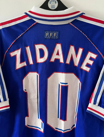 Maillot foot vintage domicile equipe de france 1998 bleu blanc et rouge. On peut retrouver l'équipementier adidas. Il s'agit d'un maillot authentique d'époque. Le maillot est floqué du numéro 10 Zinedine Zidane et comporte également sa signature avec la mention "pour toi Zinedine Zidane"