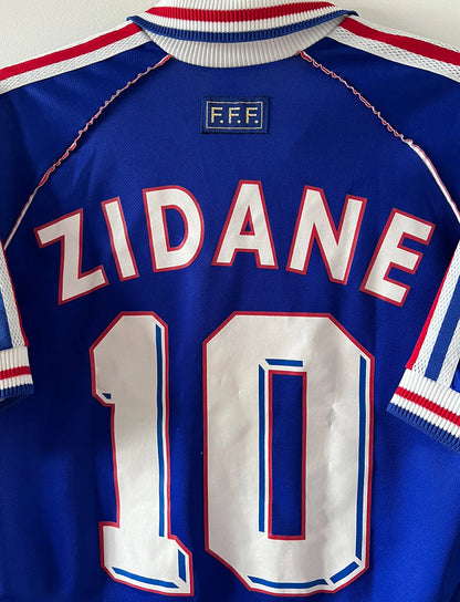 Maillot foot vintage domicile equipe de france 1998 bleu blanc et rouge. On peut retrouver l'équipementier adidas. Il s'agit d'un maillot authentique d'époque. Le maillot est floqué du numéro 10 Zinedine Zidane et comporte également sa signature avec la mention "pour toi Zinedine Zidane"