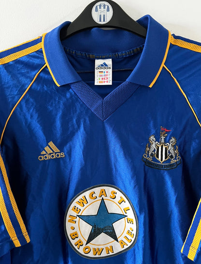 Maillot de foot vintage extérieur bleu de Newcastle 1998/1999. On peut retrouver l'équipementier Adidas et le sponsor Newcastle Brown Ale. Il s'agit d'un maillot authentique d'époque.