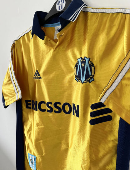 Maillot de foot vintage third de l'OM de la saison 1998-1999. Il s'agit du maillot édité pour le centenaire du club. Le maillot est de couleur dorée et bleu. On peut retrouver l'équipementier adidas et le sponsor ericsson. Il s'agit d'un maillot authentique d'époque.