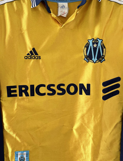 Maillot de foot vintage third de l'OM de la saison 1998-1999. Il s'agit du maillot édité pour le centenaire du club. Le maillot est de couleur dorée et bleu. On peut retrouver l'équipementier adidas et le sponsor ericsson. Il s'agit d'un maillot authentique d'époque.