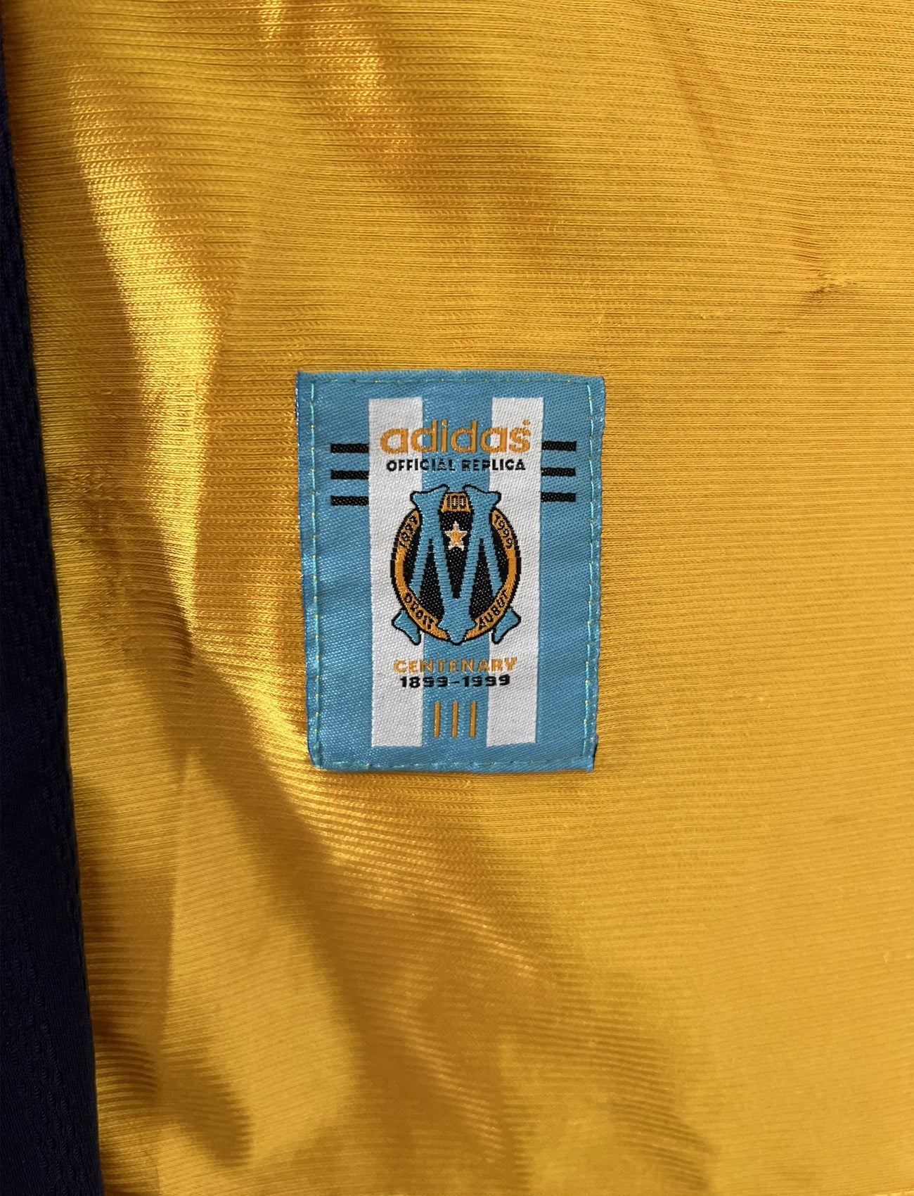 Maillot de foot vintage third de l'OM de la saison 1998-1999. Il s'agit du maillot édité pour le centenaire du club. Le maillot est de couleur dorée et bleu. On peut retrouver l'équipementier adidas et le sponsor ericsson. Il s'agit d'un maillot authentique d'époque.