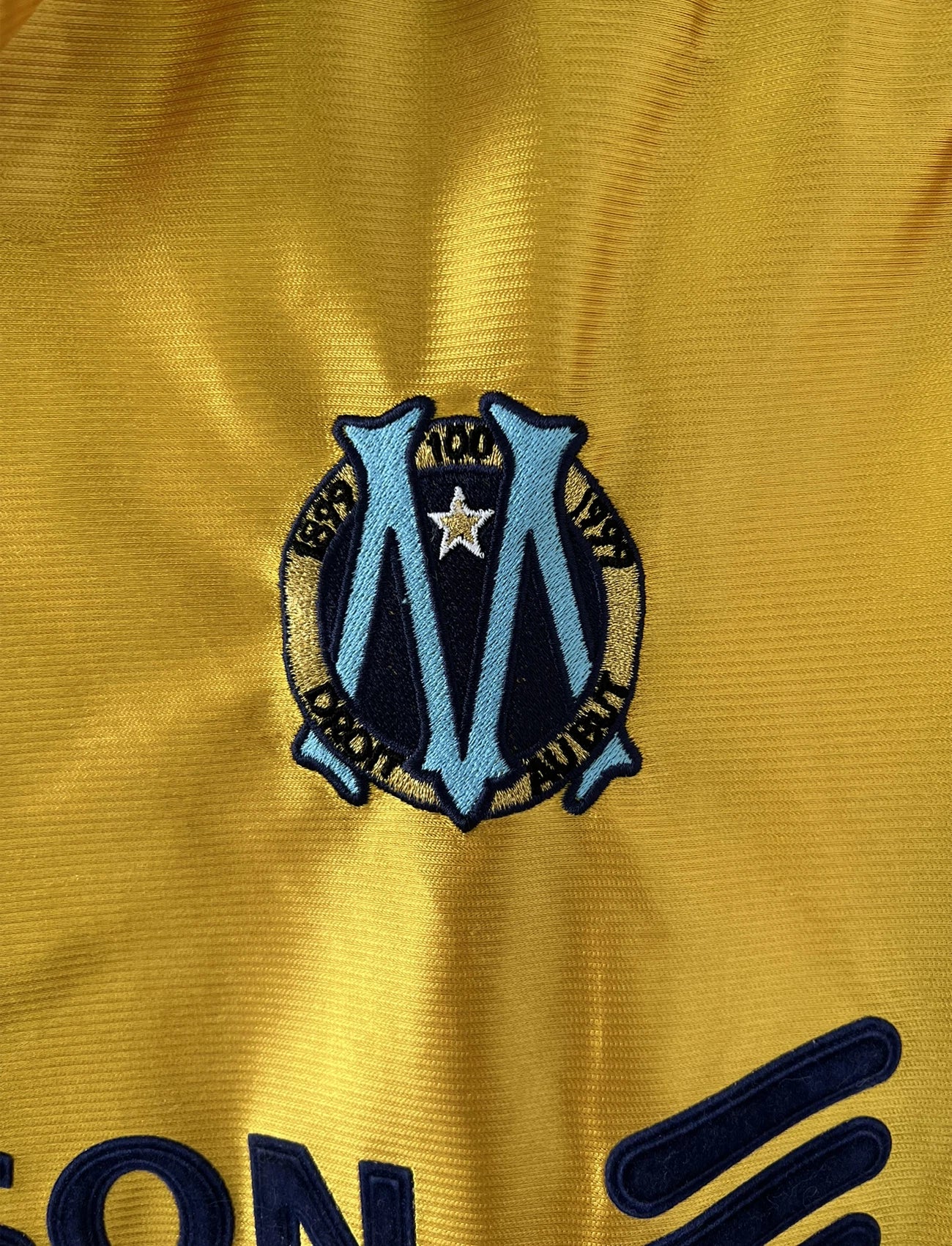 Maillot de foot vintage third de l'OM de la saison 1998-1999. Il s'agit du maillot édité pour le centenaire du club. Le maillot est de couleur dorée et bleu. On peut retrouver l'équipementier adidas et le sponsor ericsson. Il s'agit d'un maillot authentique d'époque.