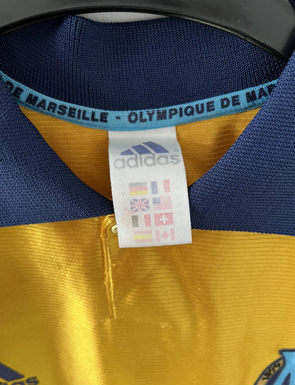 Maillot de foot vintage third de l'OM de la saison 1998-1999. Il s'agit du maillot édité pour le centenaire du club. Le maillot est de couleur dorée et bleu. On peut retrouver l'équipementier adidas et le sponsor ericsson. Il s'agit d'un maillot authentique d'époque.