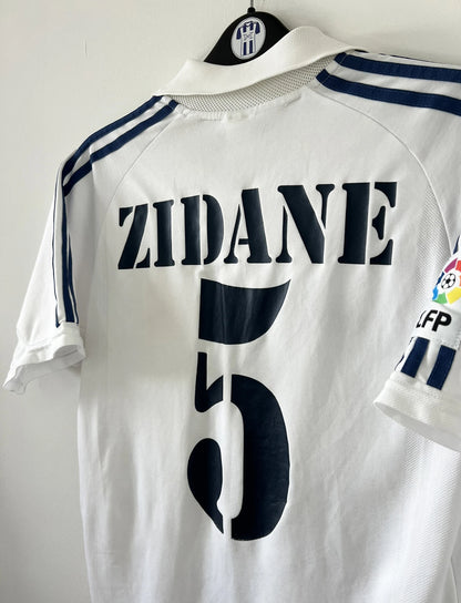 Maillot de foot vintage domicile du real madrid de la saison 2001-2002. Le maillot est de couleur blanc et noir. On peut retrouver l'équipementier adidas et le sponsor Real Madrid.com. Le maillot est floqué du numéro 5 Zinedine Zidane. Il s'agit d'un maillot authentique