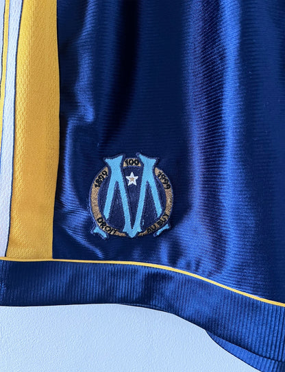 Maillot + short vintage third doré de l'OM 1998-1999. On peut retrouver l'équipementier Adidas et le sponsor Ericsson. Il s'agit d'un maillot et d'un short authentique d'époque.