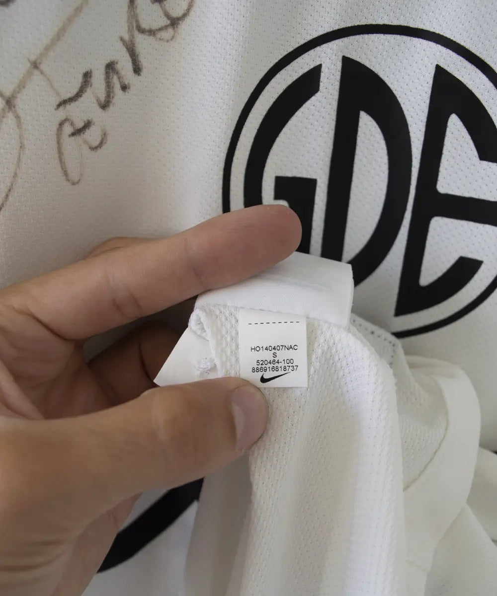 Maillot third du SM Caen de la saison 2014-2015 de couleur blanc et noir. On peut retrouver l'équipementier nike, le sponsor GDE et le sponsor SAS Malus. Le maillot est floqué du numéro 2 Nicolas Seube. Le maillot est aussi signé par Seube. Sur cette photo on peut voir l'étiquette du maillot comportant les numéros 520464-100
