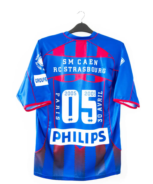 Maillot domicile du SM Caen édité lors de la finale de la coupe de la ligue face à Strasbourg. Le maillot est de couleur rouge et bleu. On peut retrouver la date de la finale, l'équipementier errea et le sponsor phillips